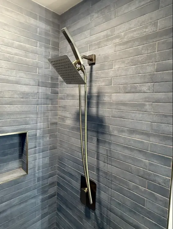 Shower wall of narrow blue-gray plank tile in an offset pattern, with a square rain head, a hand shower on a slide bar and a small niche.