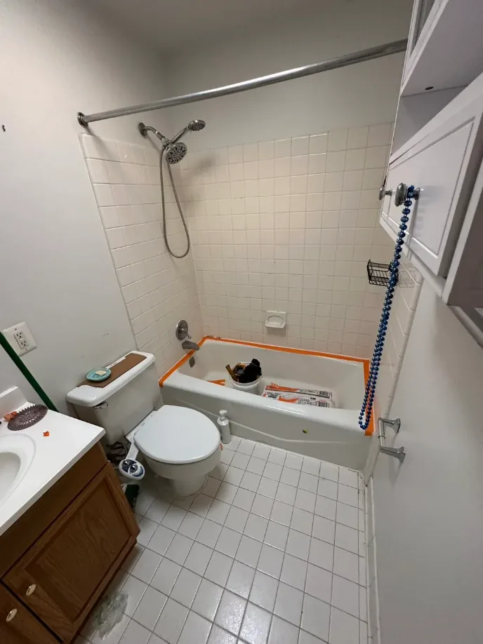 Before: hall bath with small white square floor tile, beige tile around the tub and an oak vanity.