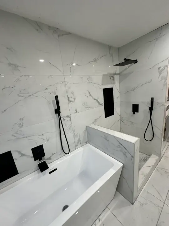 Freestanding tub beside a tiled half wall, with an open shower behind it and matte black fixtures on veined porcelain walls.